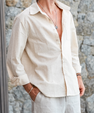 Man wearing a beige linen long sleeve casual shirt with rolled cuffs and white pants standing against stone wall